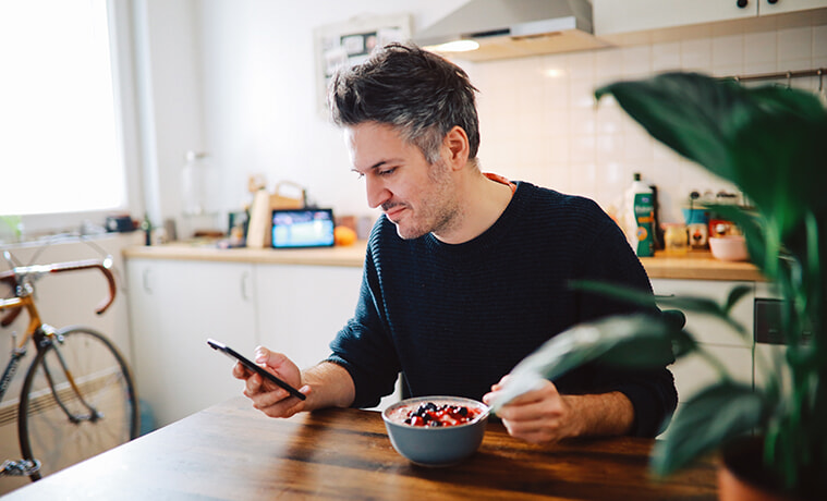 Man enjoys a healthy breakfast at his kitchen table while browsing on his smartphone.