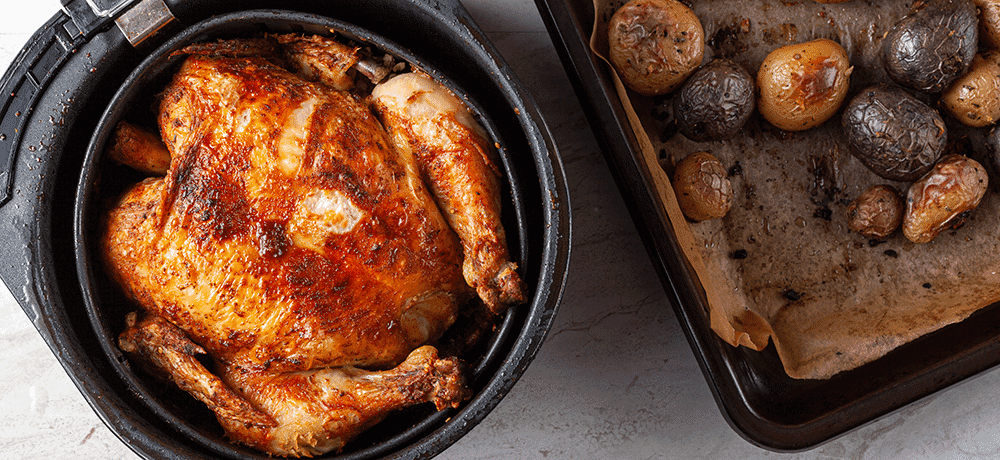A whole chicken cooked inside an air fryer with roast potatoes in an oven tray on the bench to the side