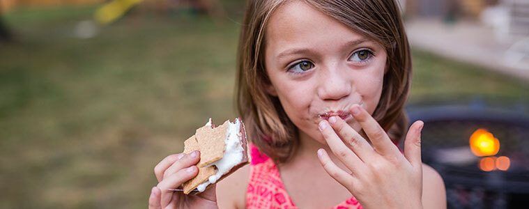 Girl looking away while eating a s’more in her yard with a barbecue grill in the background