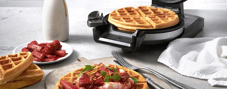 A waffle cooks inside a waffle maker, a finished waffle sits on the table covered in toppings