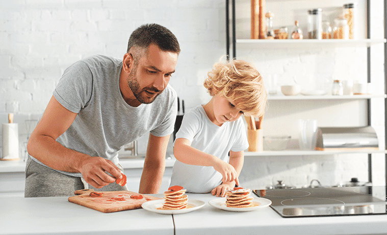 A father and son plate up stacks of pancakes with fresh strawberries at their kitchen bench