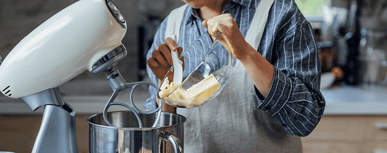 A woman adds butter to the bowl of a stand mixer while working at her kitchen bench