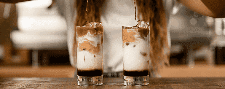 A woman pours espresso into two tall glasses of milk, to make iced coffees. 