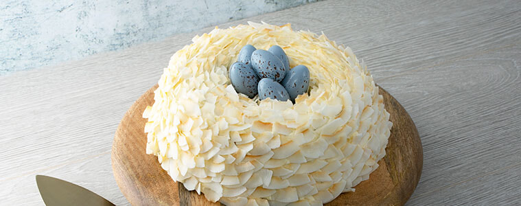 Easter Nest Cake covered in coconut flakes with blue eggs on the top, with the cake on a wooden board with a brass cake server