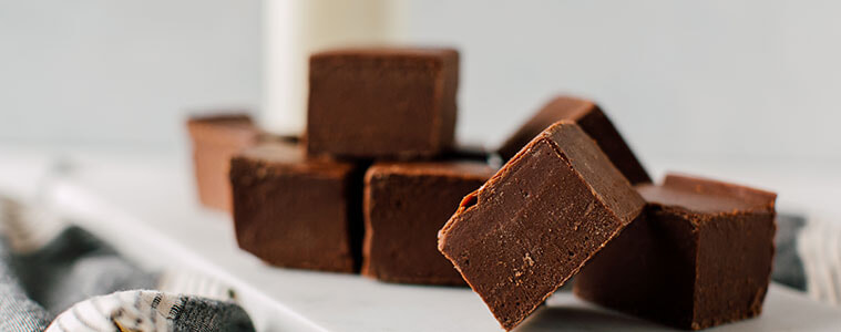 Squares of fudge, made with the Sunbeam Five-Minute fudge recipe, piled on a wooden board on a grey and white tea towel