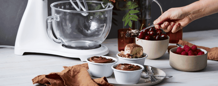 A Smeg stand mixer beside ramekins of individual Nutella puddings, fruit and chocolate sauce in a grey background and a hand holding a serving spoon