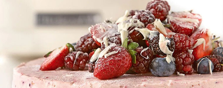 Close up image of a pink berry cheesecake, with a mound of fresh fruit berries and white chocolate shavings