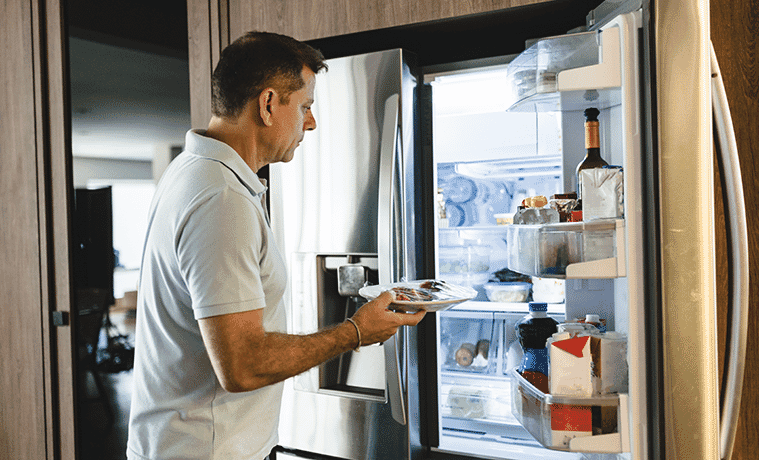 A man opens the door of the French door fridge in his kitchen to remove a plate of food