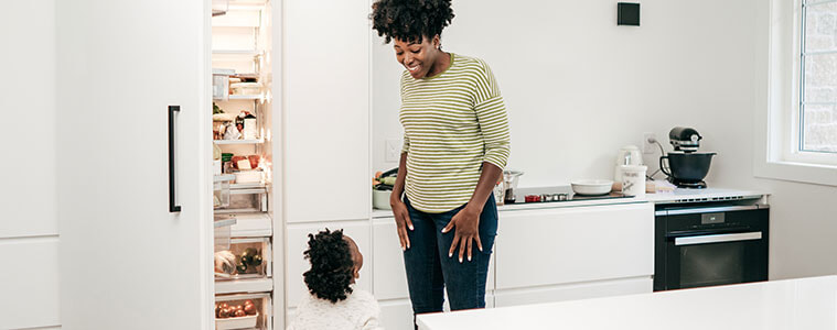 A mother and her young daughter stand next to the open door of their fridge, integrated to match white kitchen cabinetry