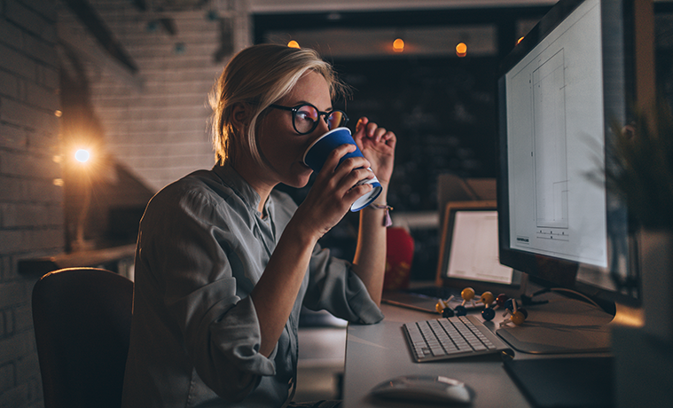 A woman sips coffee while working on a desktop computer in her home office at night