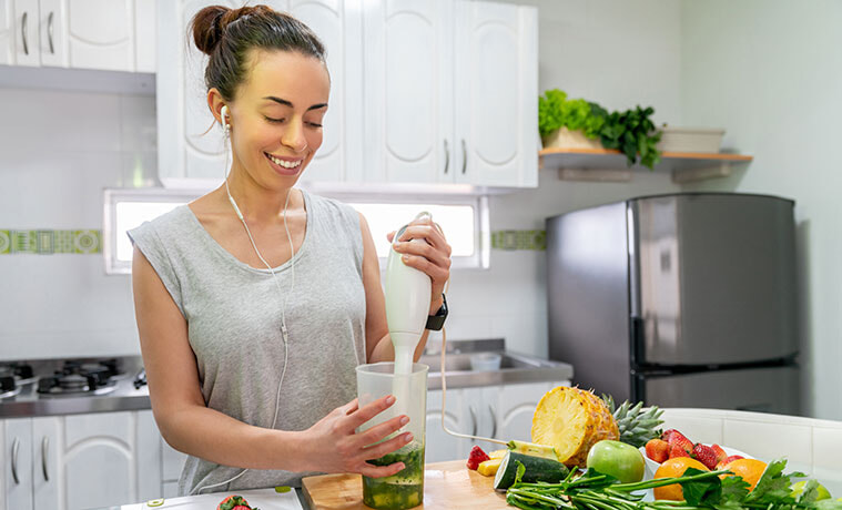 Smiling woman in workout wear making a green smoothie with a stick mixer