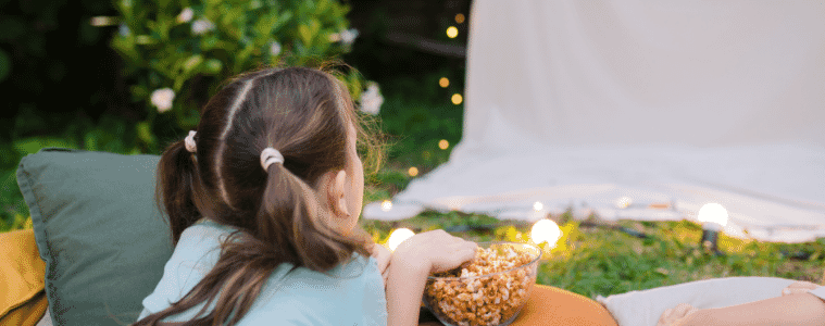 A young girl lies outside next to a bowl of popcorn, watching a movie on a projector screen