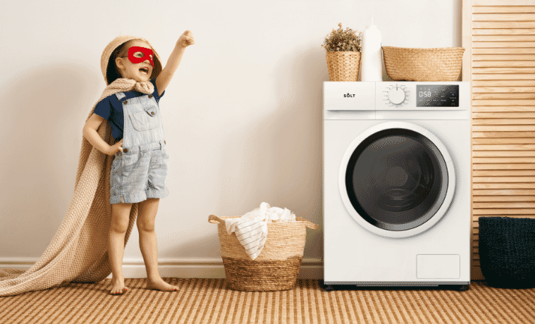 A child wears a superhero mask and uses a clean towel as a cape. He stands in a superhero pose next to a Solt washing machine.