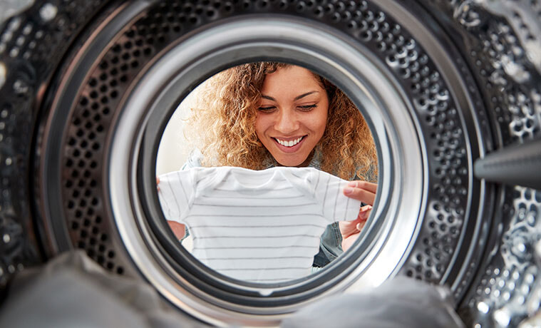 View of woman holding baby’s clothes and she leans into the open door of a tumble dryer