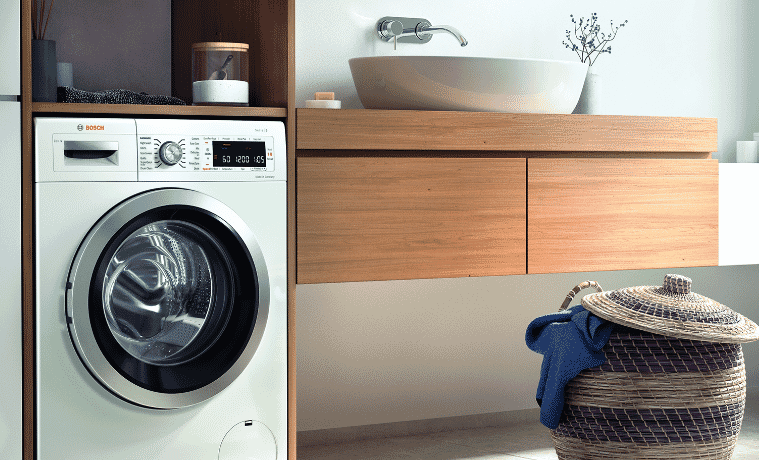 A Bosch washing machine sits next to a small basket with a blue shirt hanging over the edge of  the basket.