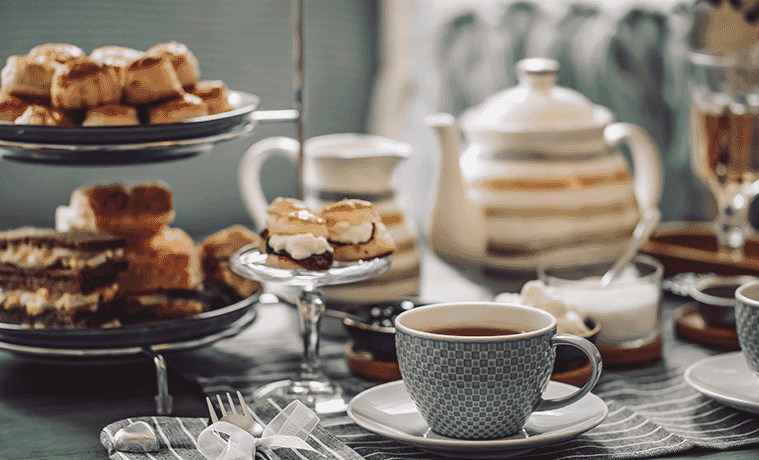 A table laid for afternoon tea with a teapot, grey china cup and saucer, cake stand topped with scones with jam and cream, and assorted cakes on a tiered cake stand
