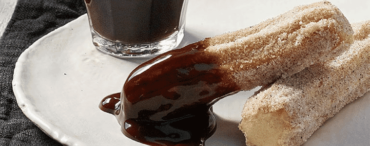 Two churros, dipped in chocolate on a white plate, sitting on a black napkin next to a cup of black coffee