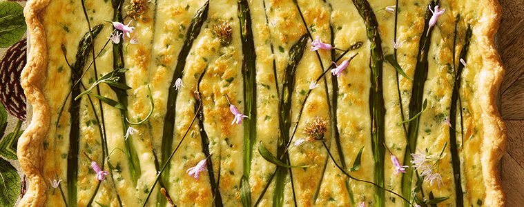 Asparagus Thyme Tart with wavy pastry edges and spears with small purple flowers on a pine cone print napkin on a wooden board