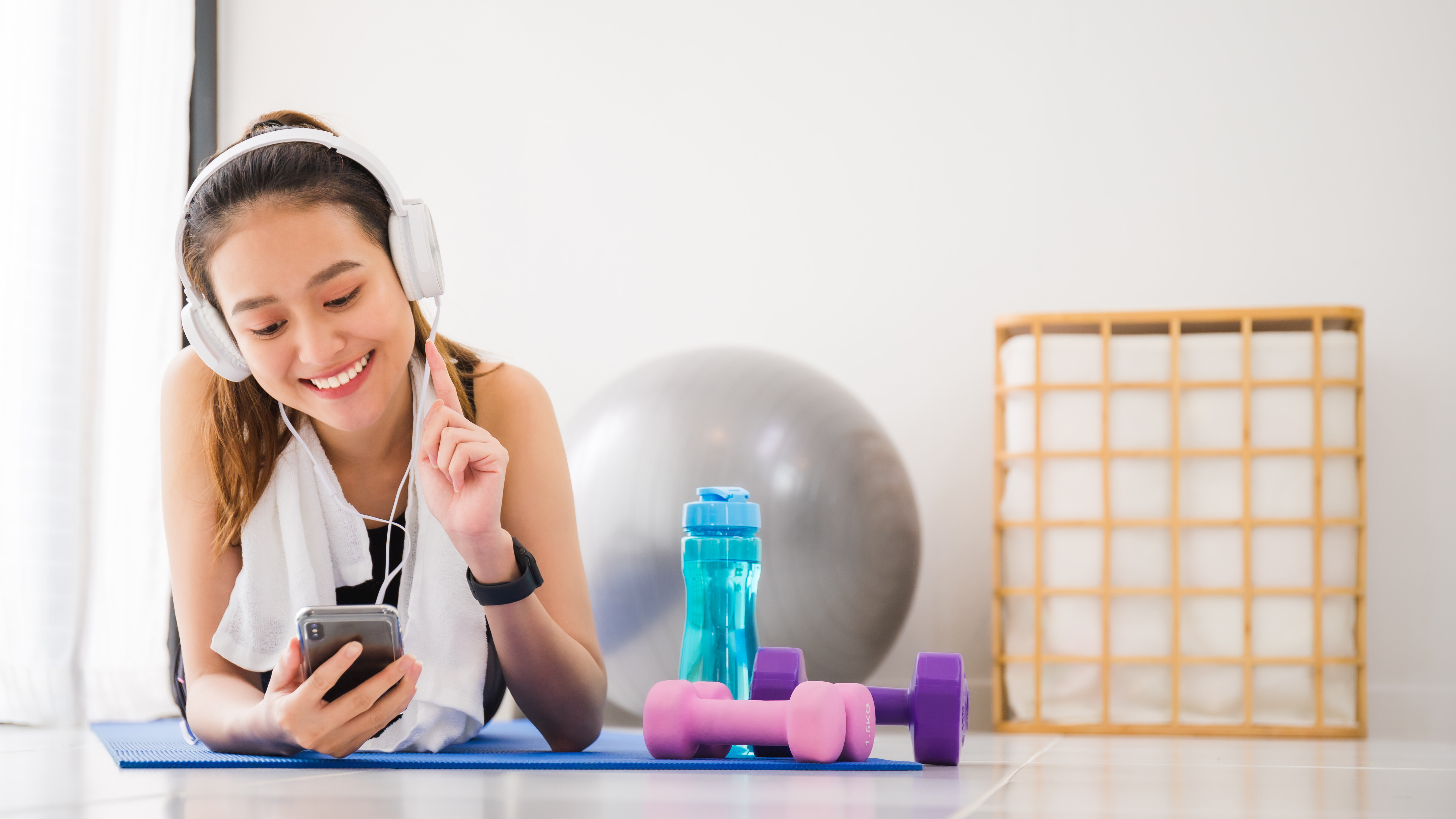 Woman listening to music with headphones and smartphone lying on yoga mat in home gym