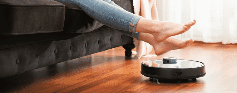 A woman sits on a couch and lifts her feet as a robot vacuum cleans under her