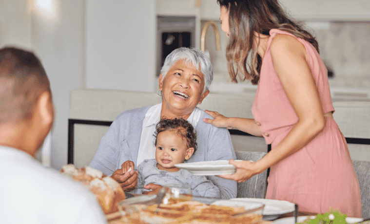 A grandmother sits at a table with a child in her lap as her adult daughter serves her a plate