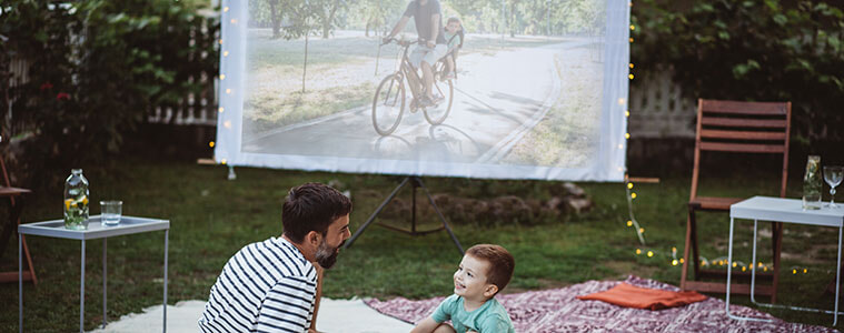 A father and son sit on a blanket together to watch a film projected onto a screen in their backyard