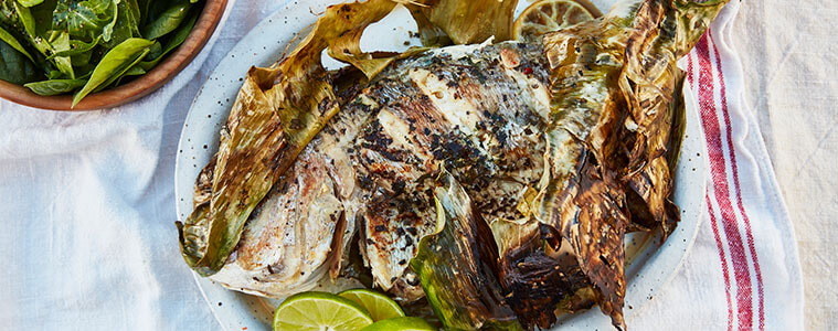 Whole grilled fish on white plate with lime slices, next to a bowl of spinach leaves, all sitting on a white teatowel