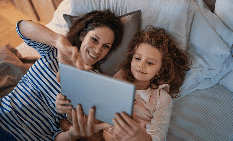 A mother and daughter lie on a bed at home and watch a TV show on a tablet