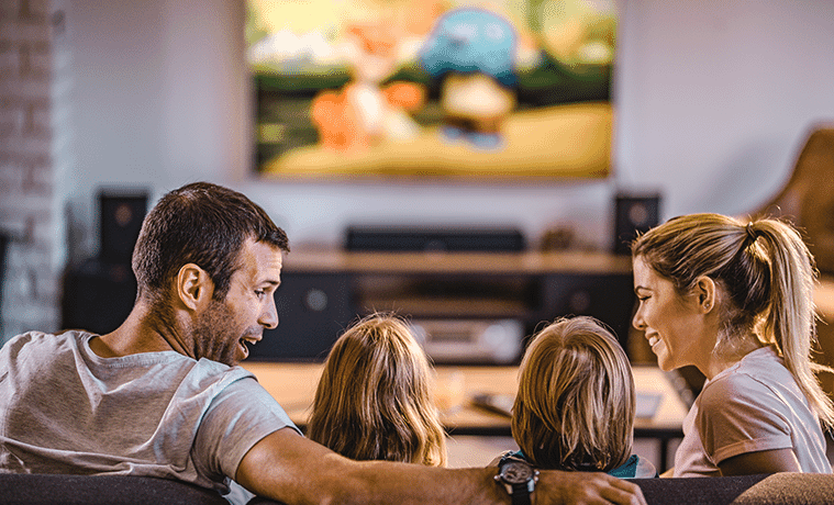 A family sits on a couch and watches a big screen TV