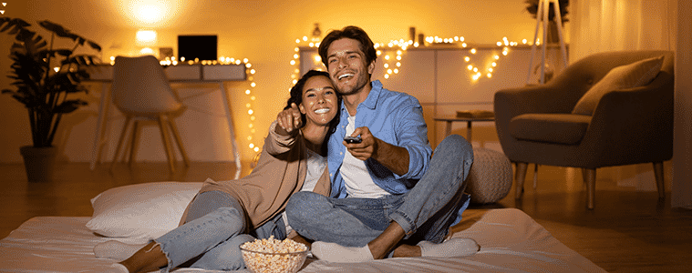 Couple sitting on their bed, watching TV, surrounded by fairy lights