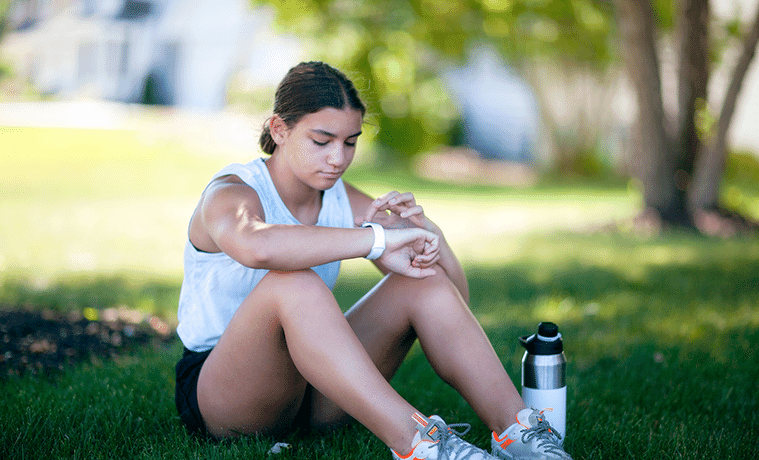 A teenage girl sits on the grass in a park to check the display on her fitness tracker