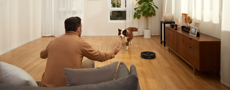 Image of a man laying with a dog in a modern living room while the Roborock S8 MaxV Ultra Robotic Vacuum works in the background.