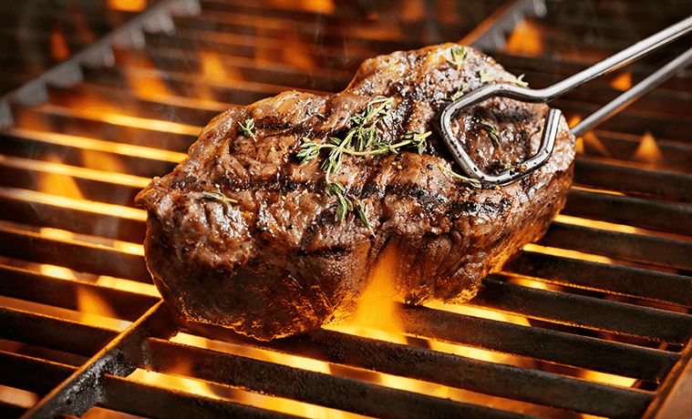 A close-up of a thick sirloin steak with fresh thyme above the flames of a barbecue grill.
