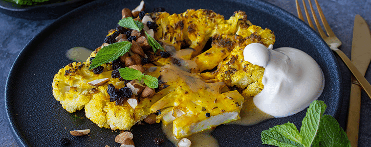 Cauliflower sliced and grilled covered with nuts, green mint and currants with a dollop of yoghurt on a black plate.