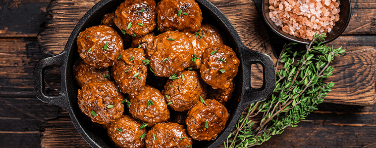 Meatballs in a cast iron serving dish on a rustic timber table with a bouquet of herbs and a bowl of pink salt flakes. 