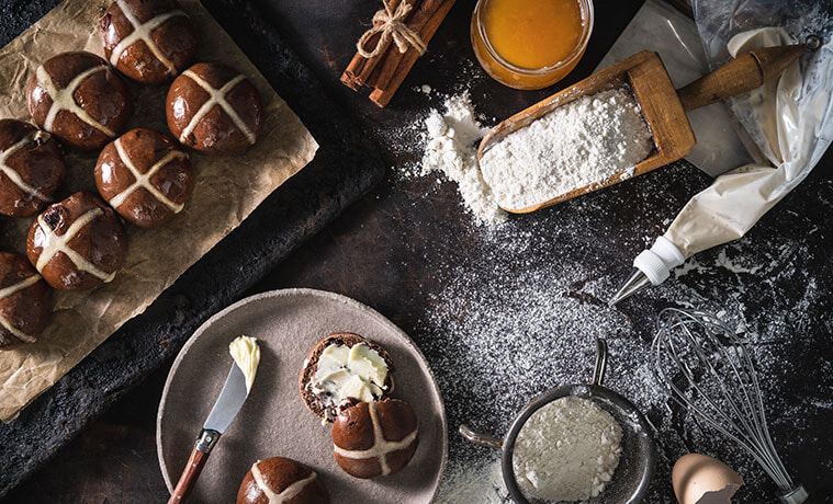Overhead view of a dark stone kitchen bench, dusted with flour, with a buttered hot cross bun on a plate, piping bag, whisk, 
