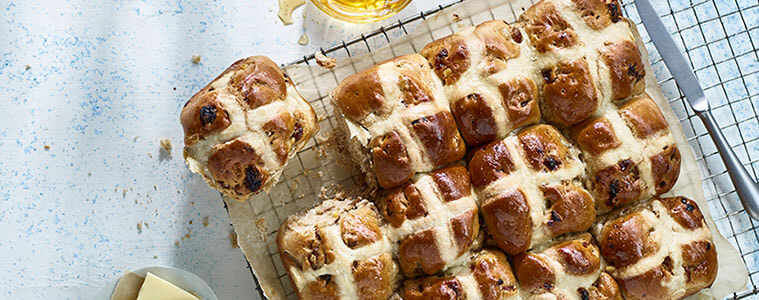 Hot cross buns on a tray lined with baking paper with mini white Easter eggs on top, sitting on a timber table with a cup of tea.