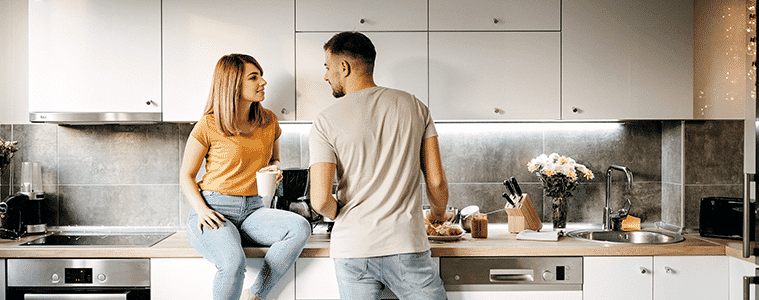 Woman sits on the kitchen bench to talk to her partner while he makes lunch in their modern white galley kitchen with an under bench oven and sleek electric cooktop. 