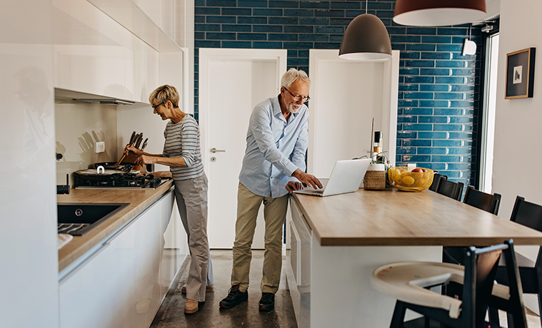 Senior couple cook together in their kitchen with white high-gloss cabinetry, stone benches and high-end kitchen appliances.