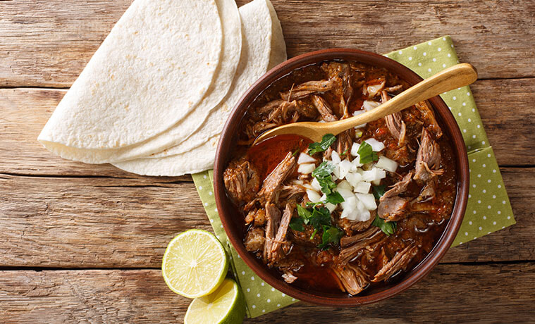 Soft tacos fanned on a timber table next to a ceramic bowl filled with slow-cooked beef and a lime wedge.