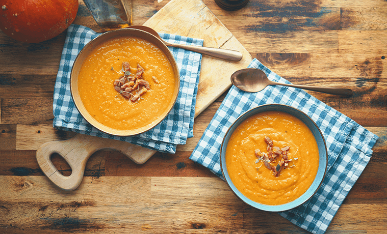 Two bowls of creamy vegetable soup sitting on blue gingham napkins on top of a timber cutting board and timber table.