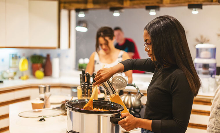 Young woman in a modern kitchen stirring a hot dish in a slow cooker, with friends in the background helping with meal preparation.