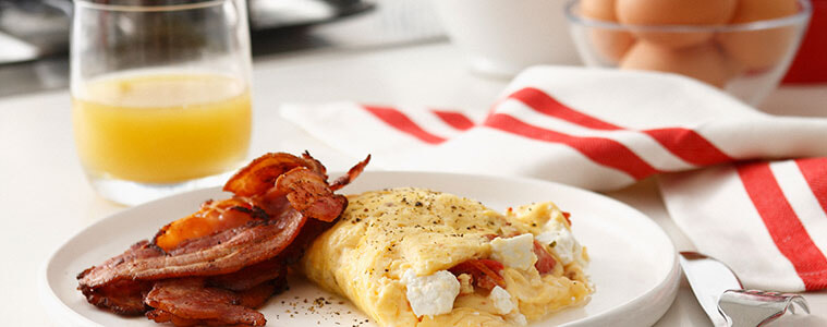 Bacon and omelette on a white plate with eggs, orange juice and frypan and cooktop in the background