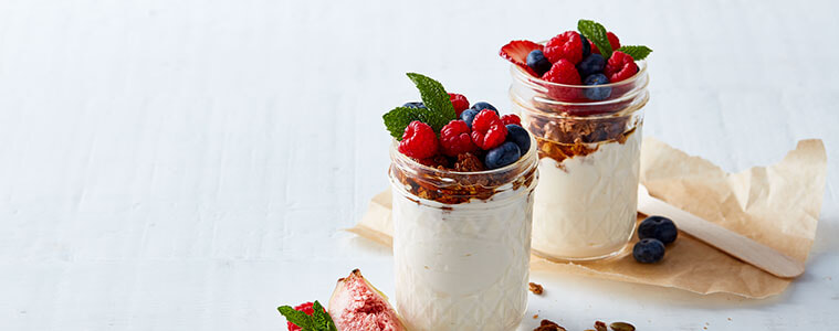 Creamy yoghurt in patterned glass jars topped with granola and fresh raspberries and blueberries on a white surface. Image and recipe by Sunbeam.