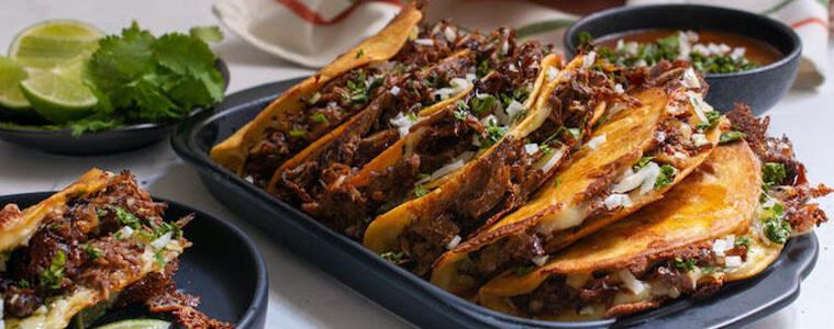 A black oven dish filled with freshly-cooked birria tacos with a plate of fresh lime wedges and coriander and a red striped tea towel in the background. Image courtesy of Weber. 