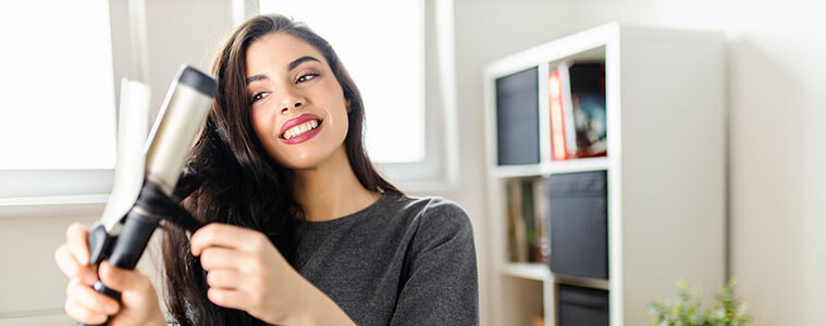 Woman using a curling iron on her long dark hair while sitting at her dressing table.