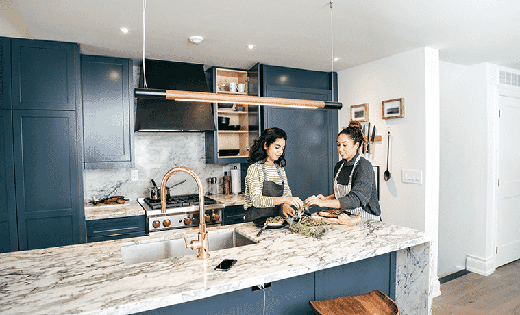 Two friends prepare dinner in a high-end kitchen with marble benchtops, blue cabinetry and brass taps.