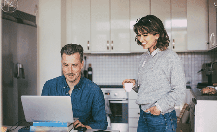 Smiling man and woman in the kitchen at home with man at the table working on a laptop.