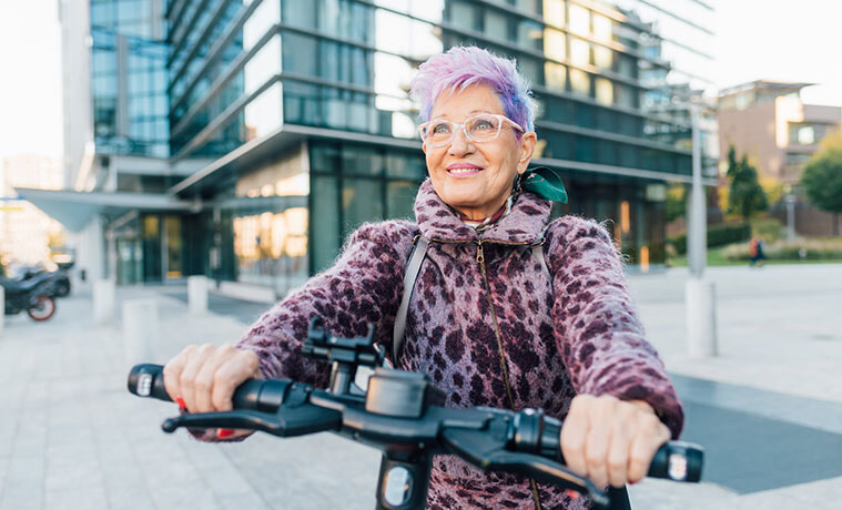 A fashionable older woman smiles while riding an e-scooter in an inner city location. 