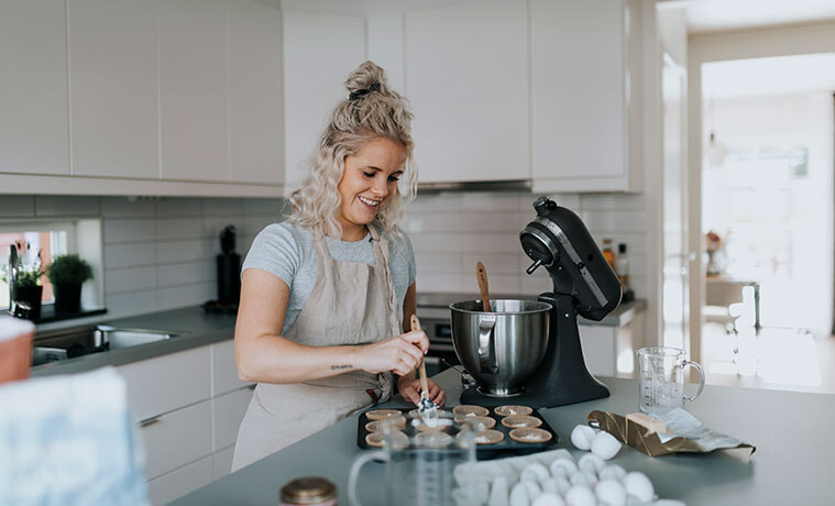Woman uses a black stand mixer to prepare cake mix for cupcakes at her kitchen bench.